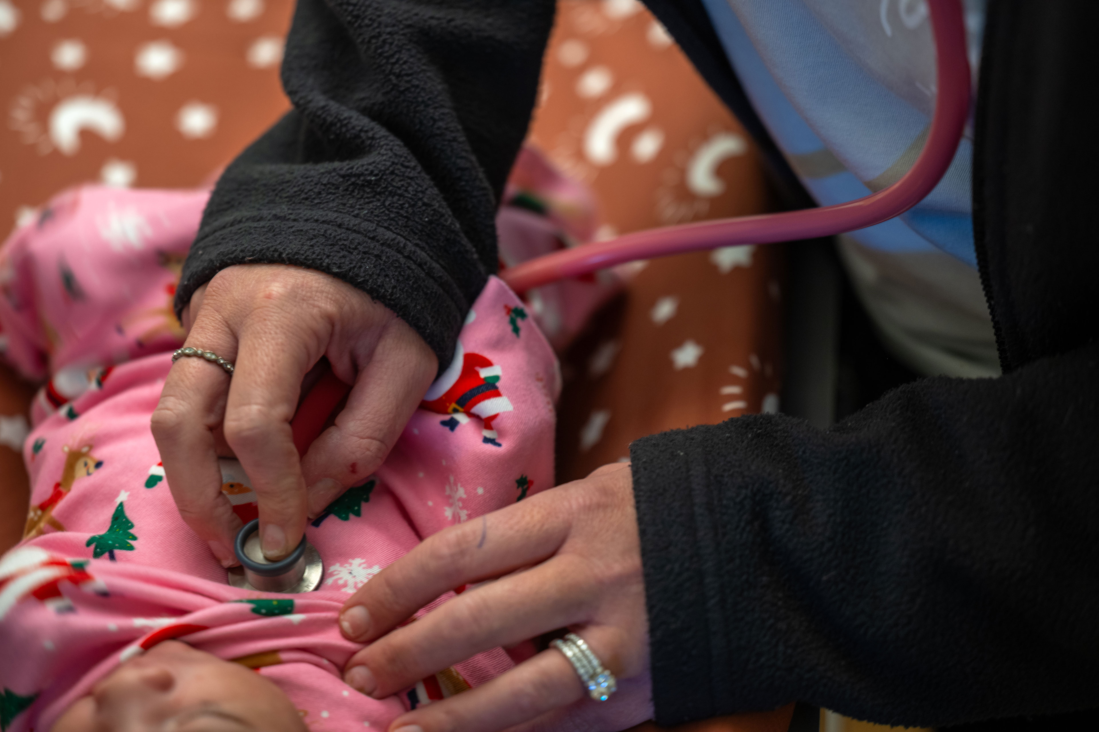 A close-up image of a nurse holding a stethoscope to baby's chest. The unidentifiable baby is wrapped in a pink cloth with a pattern of Santa, reindeer, trees, and snowflakes.