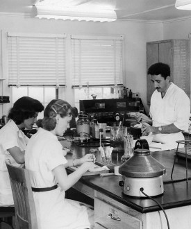 A black and white photograph from 1952 that shows four lab technicians, two men and two women, working at a table with science equipment.