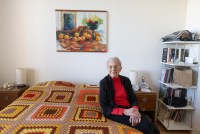 A senior woman wearing a red turtleneck and black cardigan sits on the side of her bed for a portrait. The woven blanket on her bed is made of orange, yellow, and red yarn, which matches the painting of oranges that hangs above her bed.